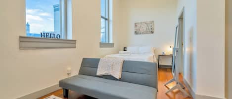 A bedroom with a bed, a grey sofa, a window, and a painting on the wall.