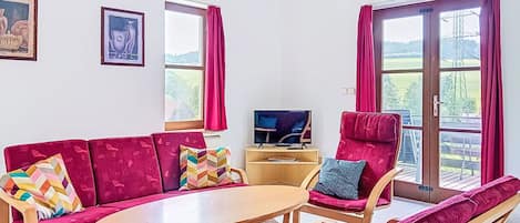 Ein Wohnzimmer mit einer Couch, Sesseln, einem Tisch, einem Fernseher und großen Fenstern mit Vorhängen.