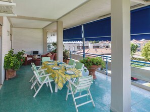 Un área de comedor al aire libre cubierta con una mesa dispuesta para cuatro personas, rodeada de sillas blancas, plantas en macetas y vistas a edificios al fondo.