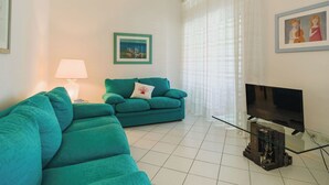Ein Wohnzimmer mit einem petrolfarbenen Sofa, einem Glastisch, einem Flachbildfernseher und einem gerahmten Bild an der Wand.