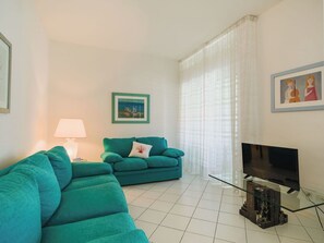 Ein Wohnzimmer mit einem petrolfarbenen Sofa, einem Glastisch, einem Flachbildfernseher und einem gerahmten Bild an der Wand.
