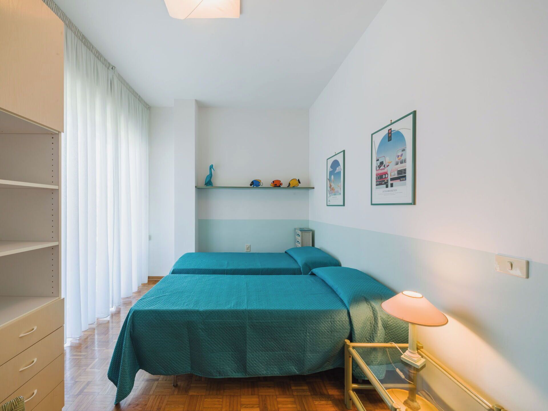 Quarto com duas camas, uma mesa de cabeceira com luminária, uma estante com objetos decorativos e um guarda-roupa.