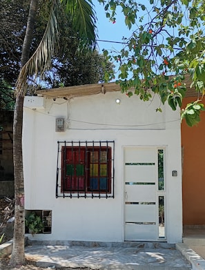 Un edificio pequeño de un piso con fachada blanca, una ventana con barrotes y una puerta con panel de vidrio.