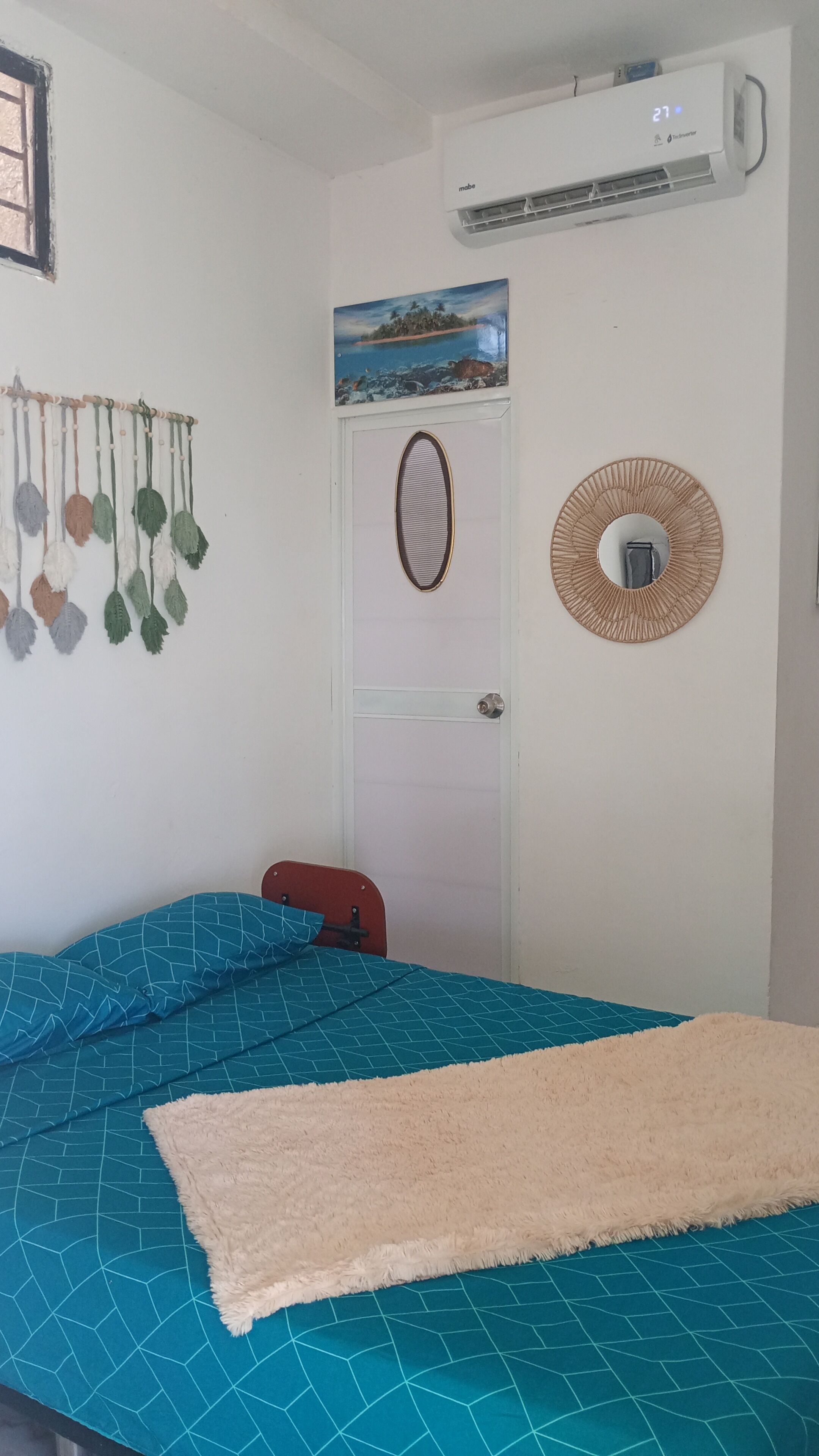 A bedroom with a bed, a wall-mounted air conditioner, a door with a decorative mirror, and a wall hanging.