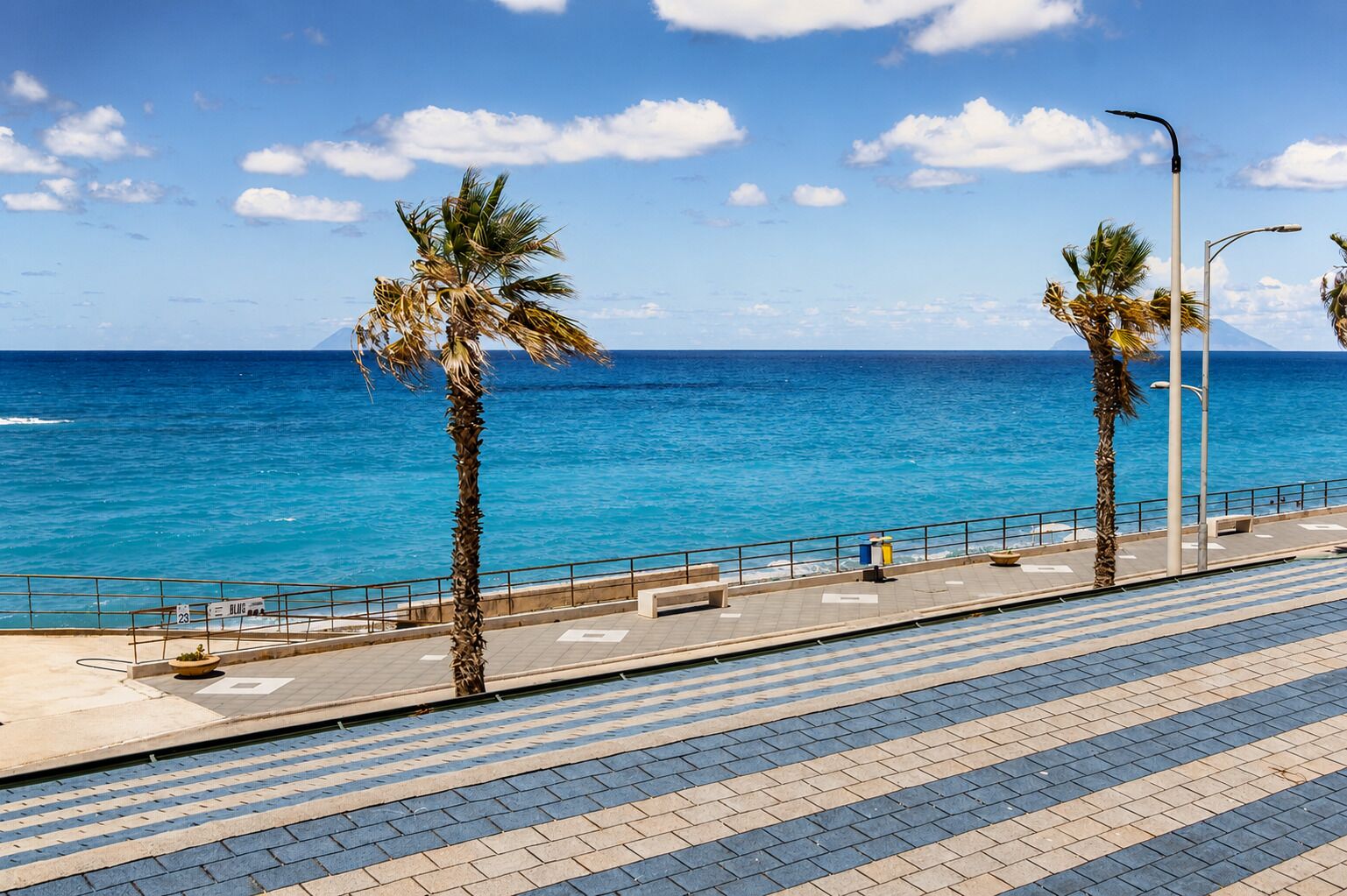 A coastal promenade with palm trees, a clear blue sky, and the ocean in the background.