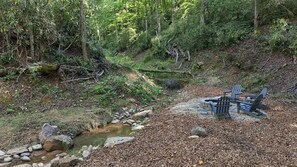 A forested area with a small stream, rocks, and a fire pit with chairs. - Mountain Thyme-Ideal location: close, with privacy, forest stream & waterfall (Saluda)