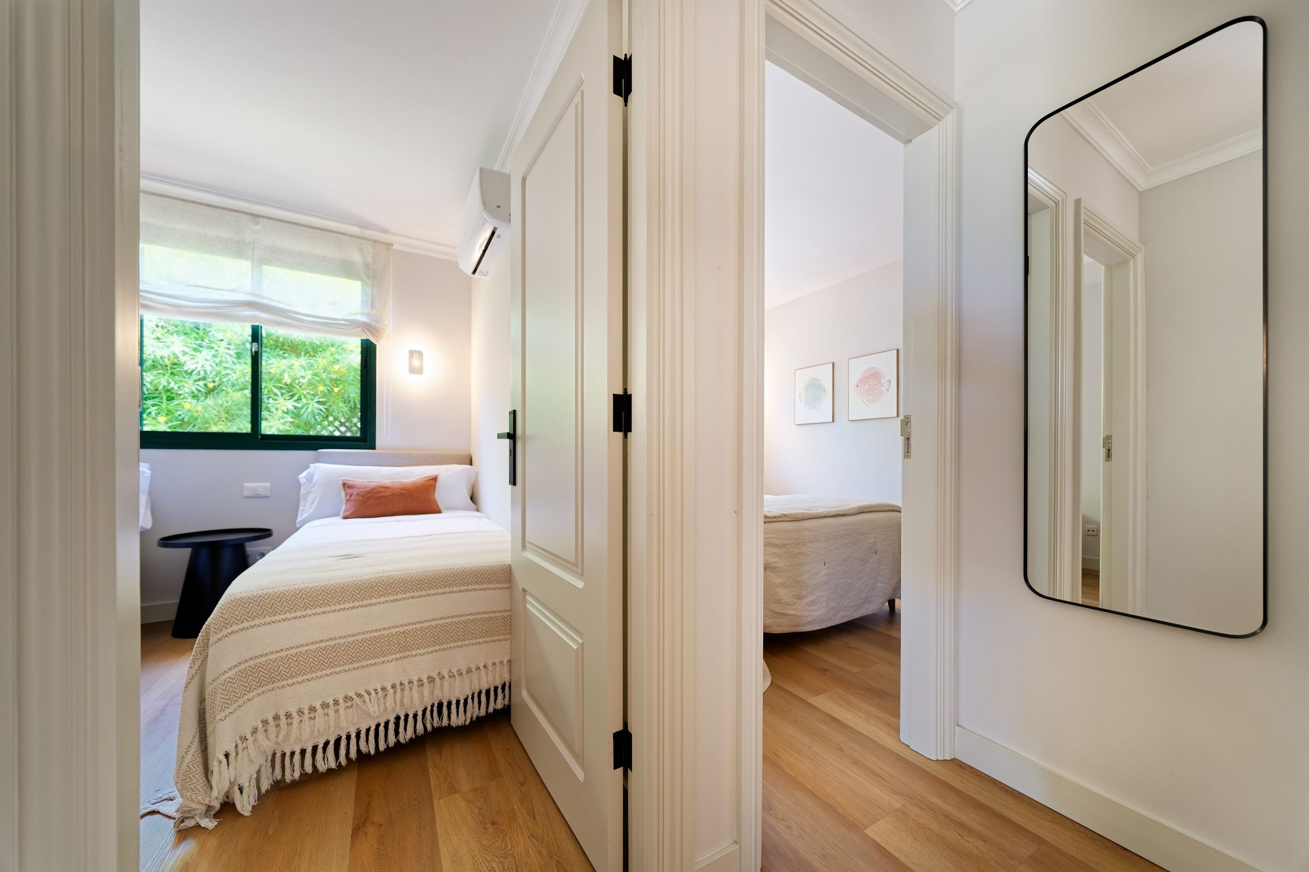 Een slaapkamer met een bed, een nachtkastje, een raam met uitzicht op groen, en een grote spiegel aan de muur.
