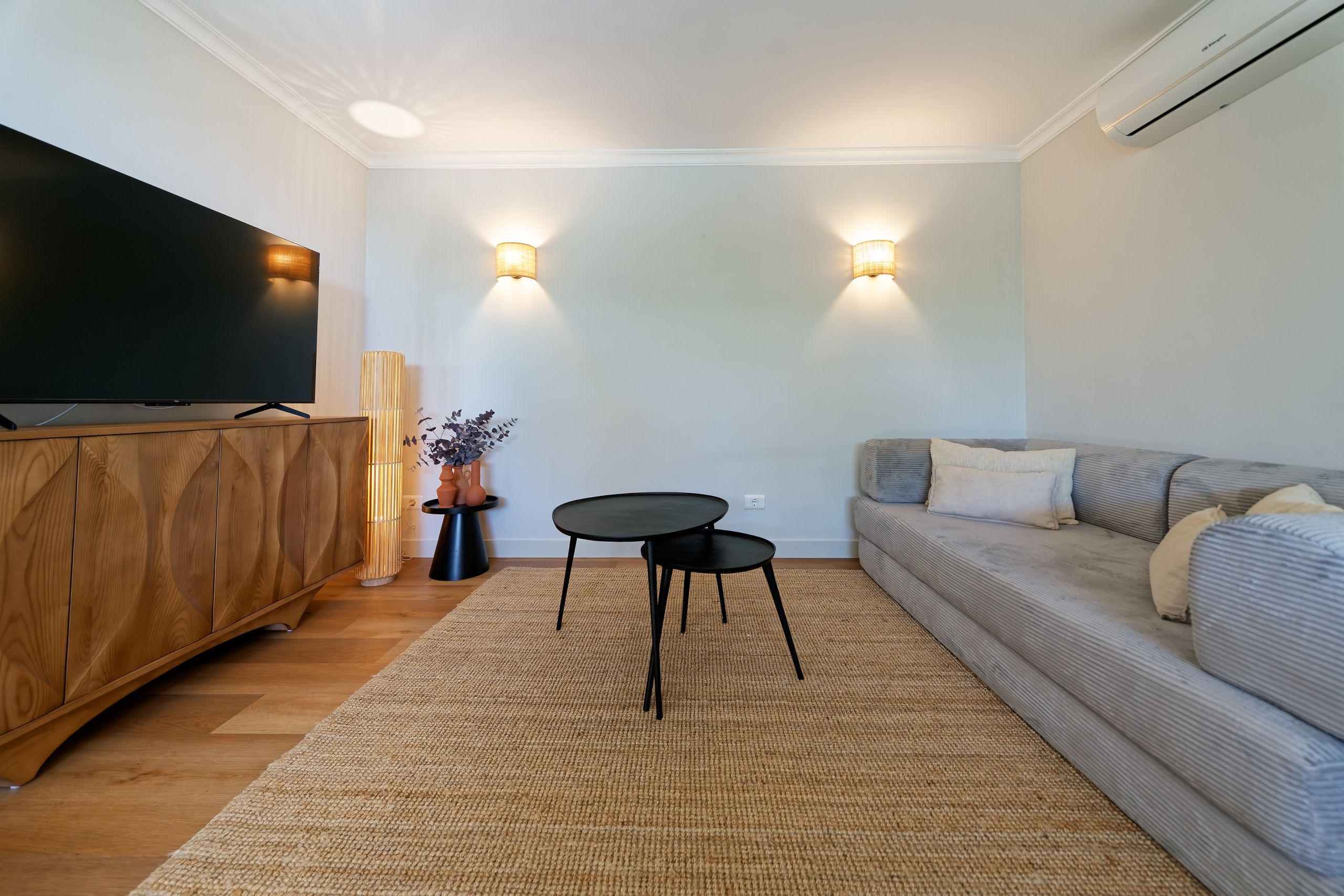 Un salon moderne avec un téléviseur à écran plat, un meuble en bois, une table ronde noire, un canapé gris et deux lampes fixées au mur.