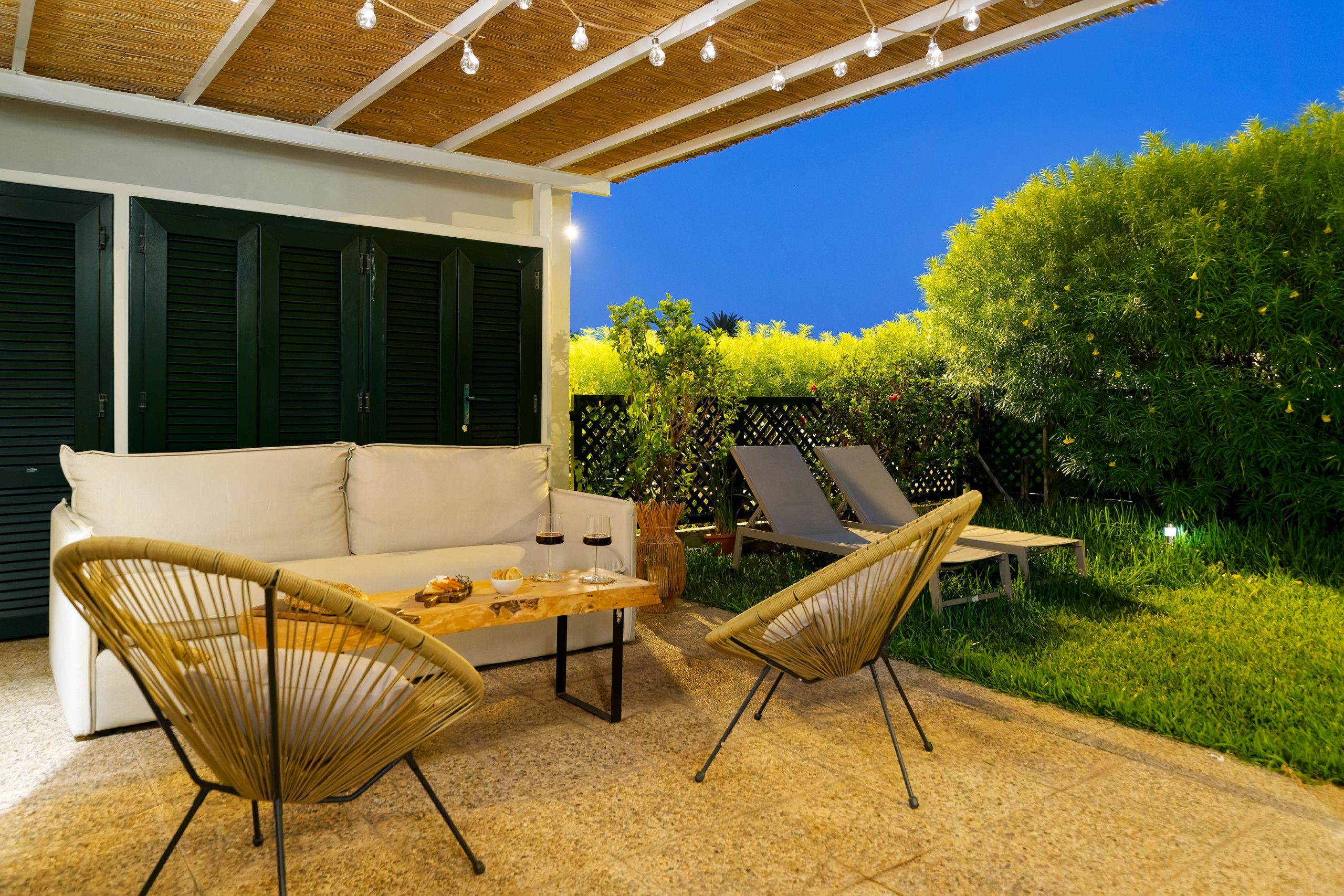 Un espace extérieur aménagé avec un canapé et deux fauteuils, une table avec des verres et un saladier, et un jardin avec une clôture et de la verdure.
