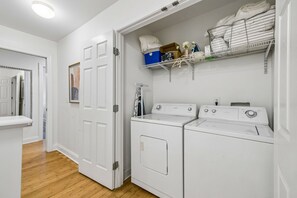 A laundry room with two white front-loading washers and dryers, a metal shelf above with folded towels, and a door leading to another room. - Spacious Nashville townhouse w/ exclusive backyard (Nashville)