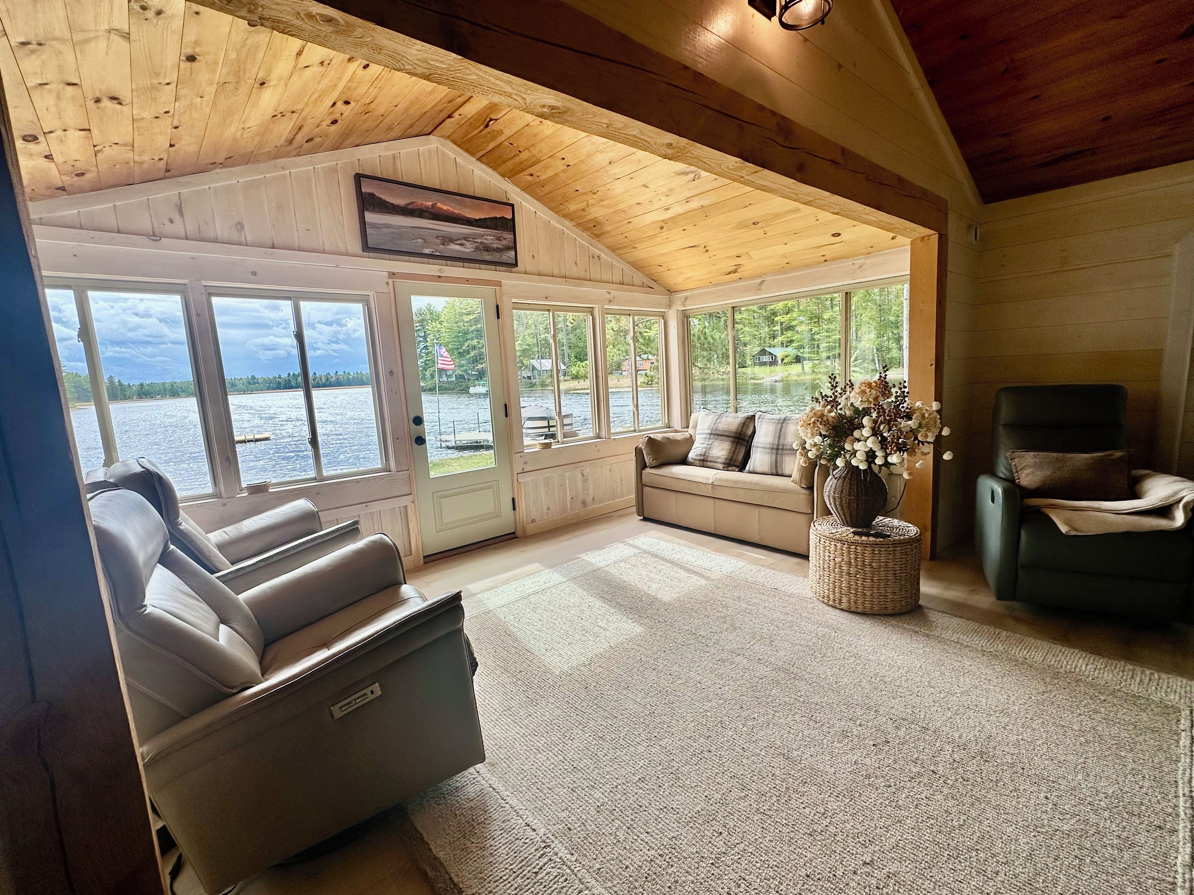 Interior acolhedor de uma cabana com uma janela grande de frente para um lago, um sofá, uma poltrona e uma mesinha com um vaso.