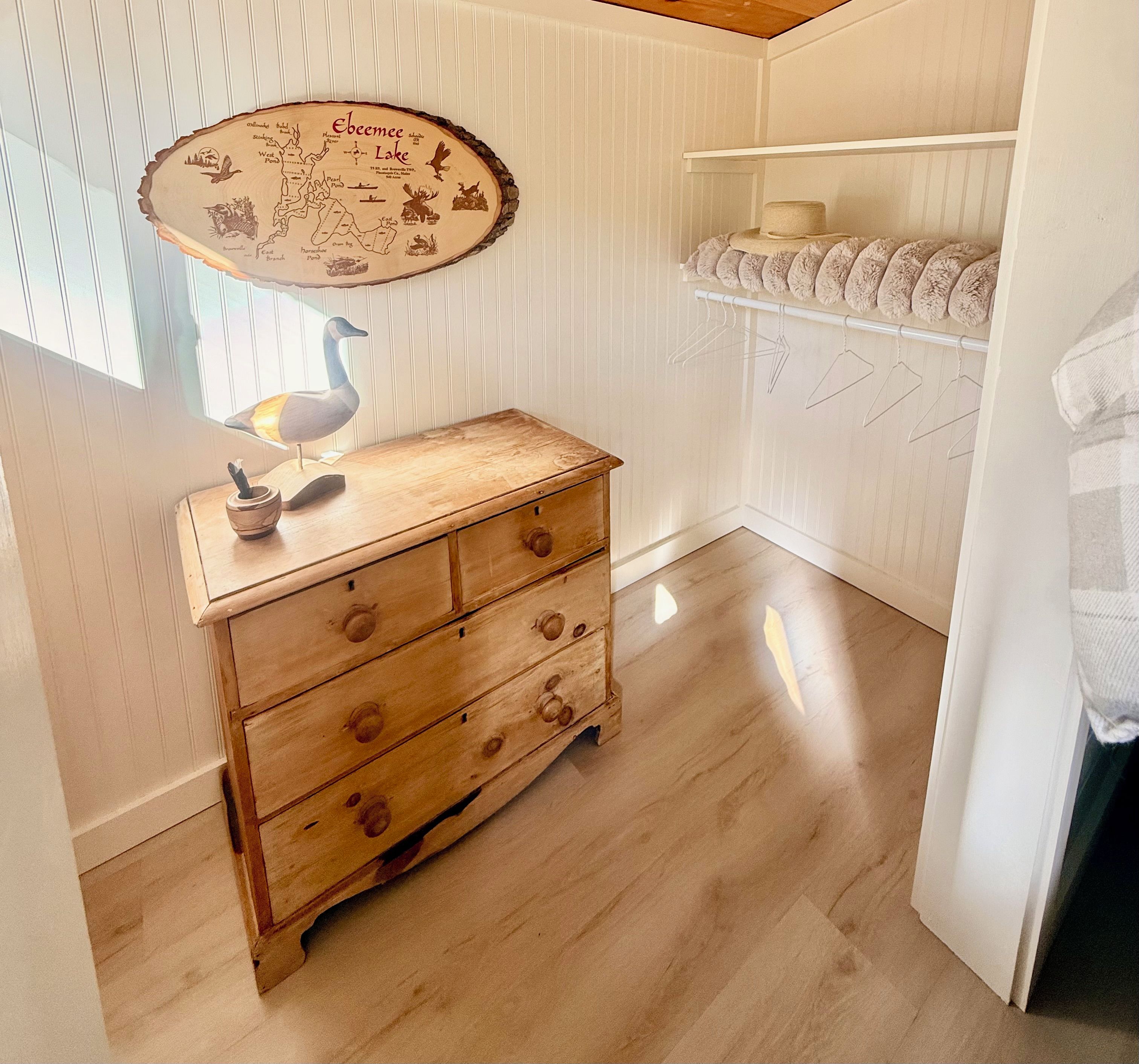 Une commode en bois avec des tiroirs, une sculpture d’oiseau en bois et une carte du lac Ebeemee accrochée au mur.