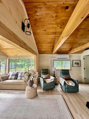 A cabin interior with a wooden ceiling, two reclining chairs, a sofa, and a large window offering a view of a lake. - Private Island Cottage on Ebeemee Lake (Brownville)