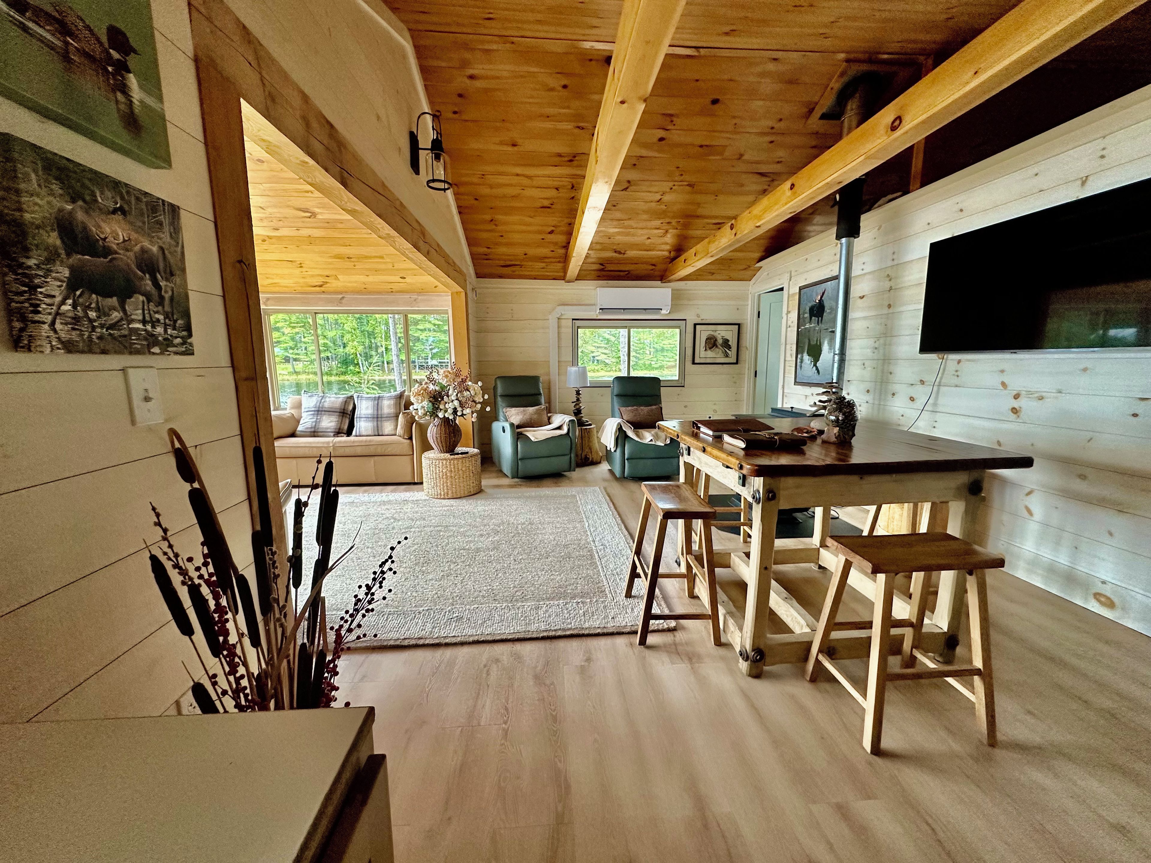 A cozy cabin interior with a wooden ceiling, a dining table with stools, a sofa, and a television.
