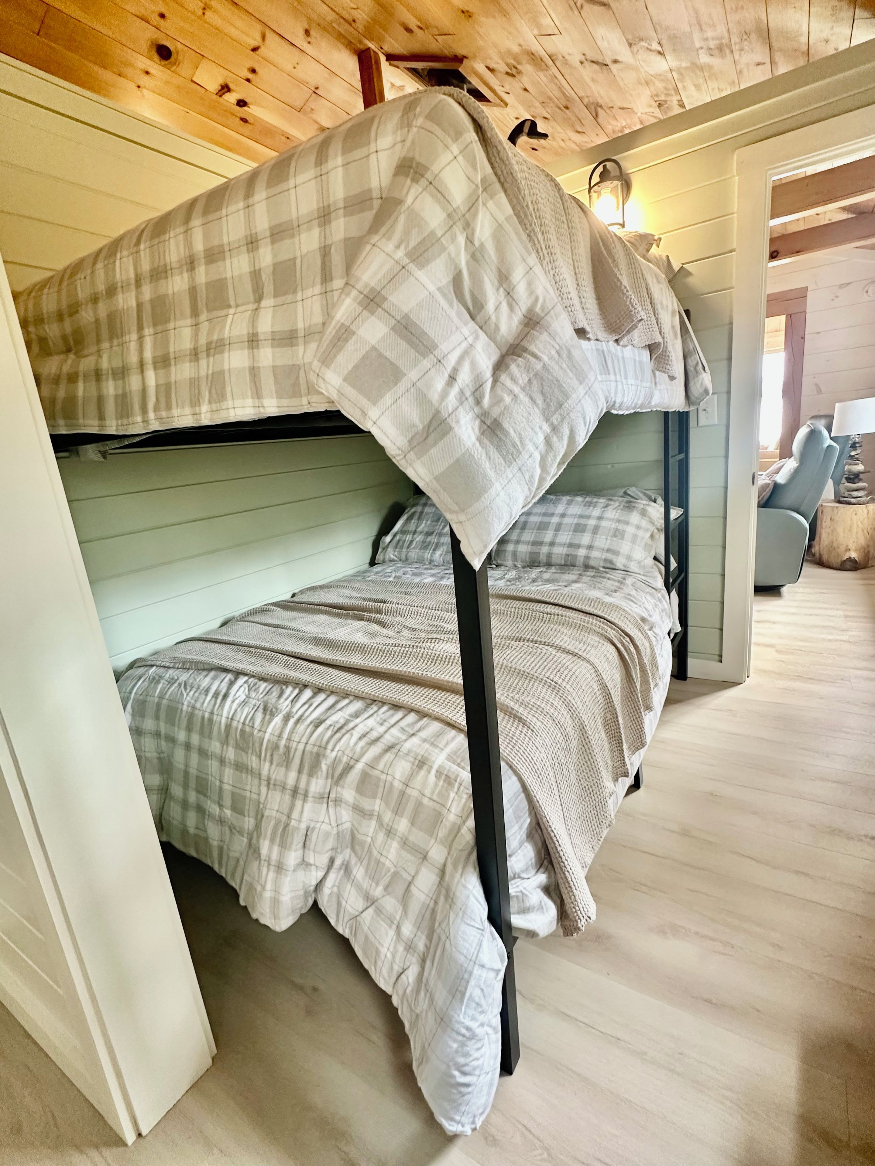 A bunk bed with a plaid blanket, a wooden floor, and a lamp on a side table.