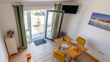 A dining area with a table set for two, a television mounted on the wall, and a view of the outdoors through a sliding glass door.