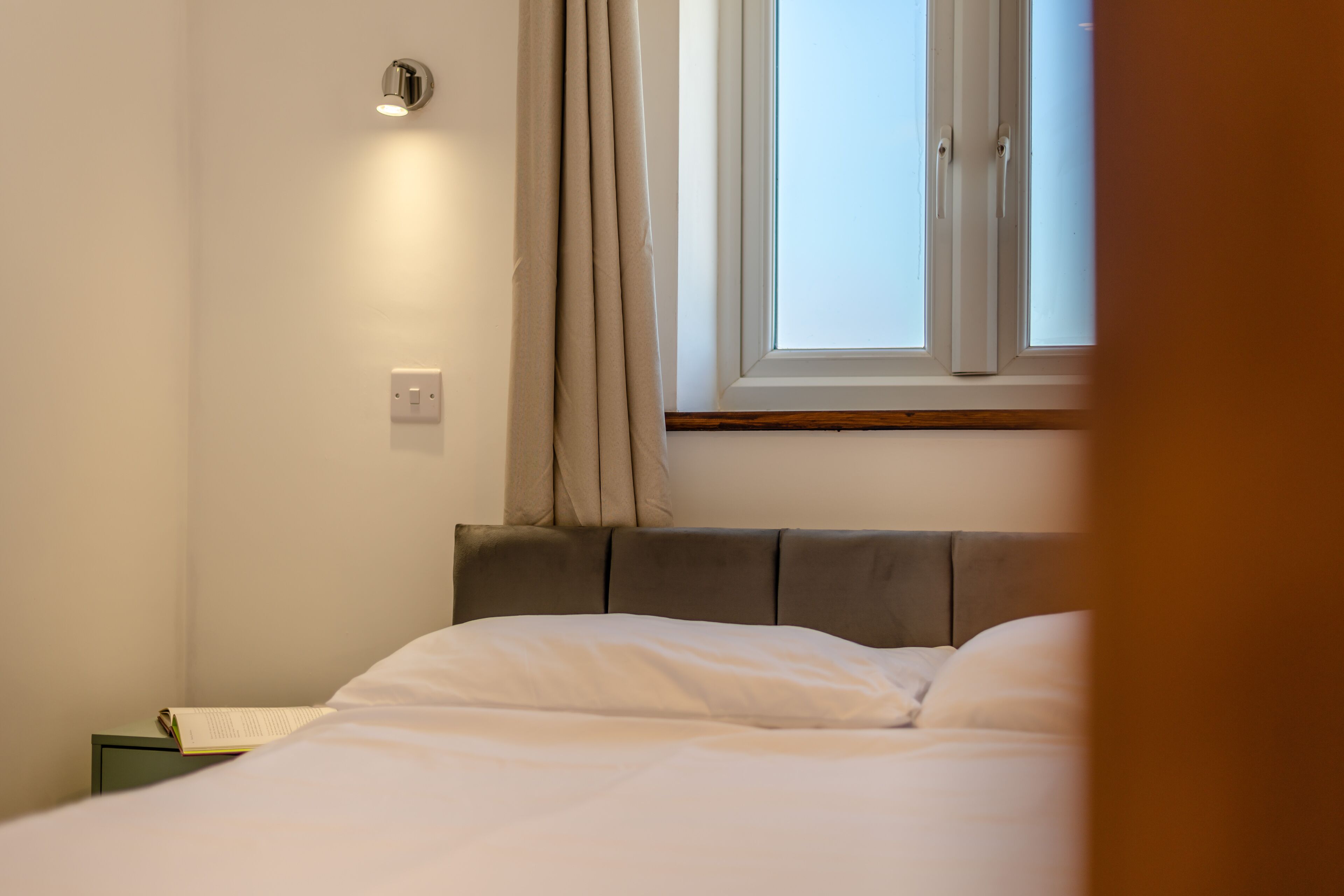 Een slaapkamer met een bed, een raam met gordijnen, een nachtkastje en een wandlamp.