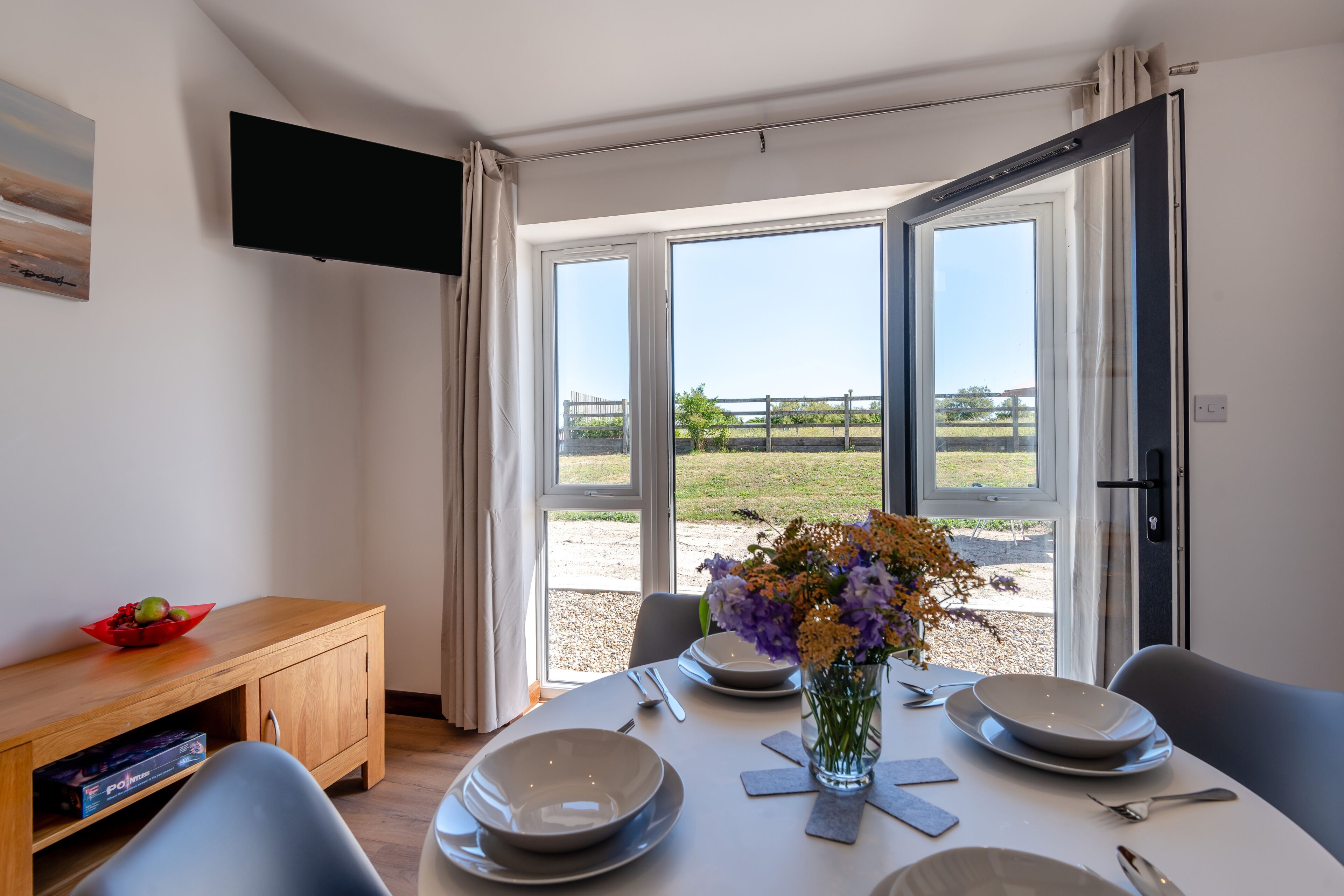 A dining area with a table set for four, a vase with flowers, and a view of a grassy field through large windows.
