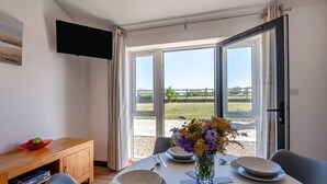 Un comedor con una mesa dispuesta para cuatro, un florero con flores y vistas a un campo cubierto de hierba a través de amplios ventanales.