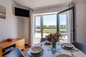 A dining area with a table set for four, a vase with flowers, and a view of a grassy field through large windows. - Kent Coast Stays (Birchington-on-Sea)