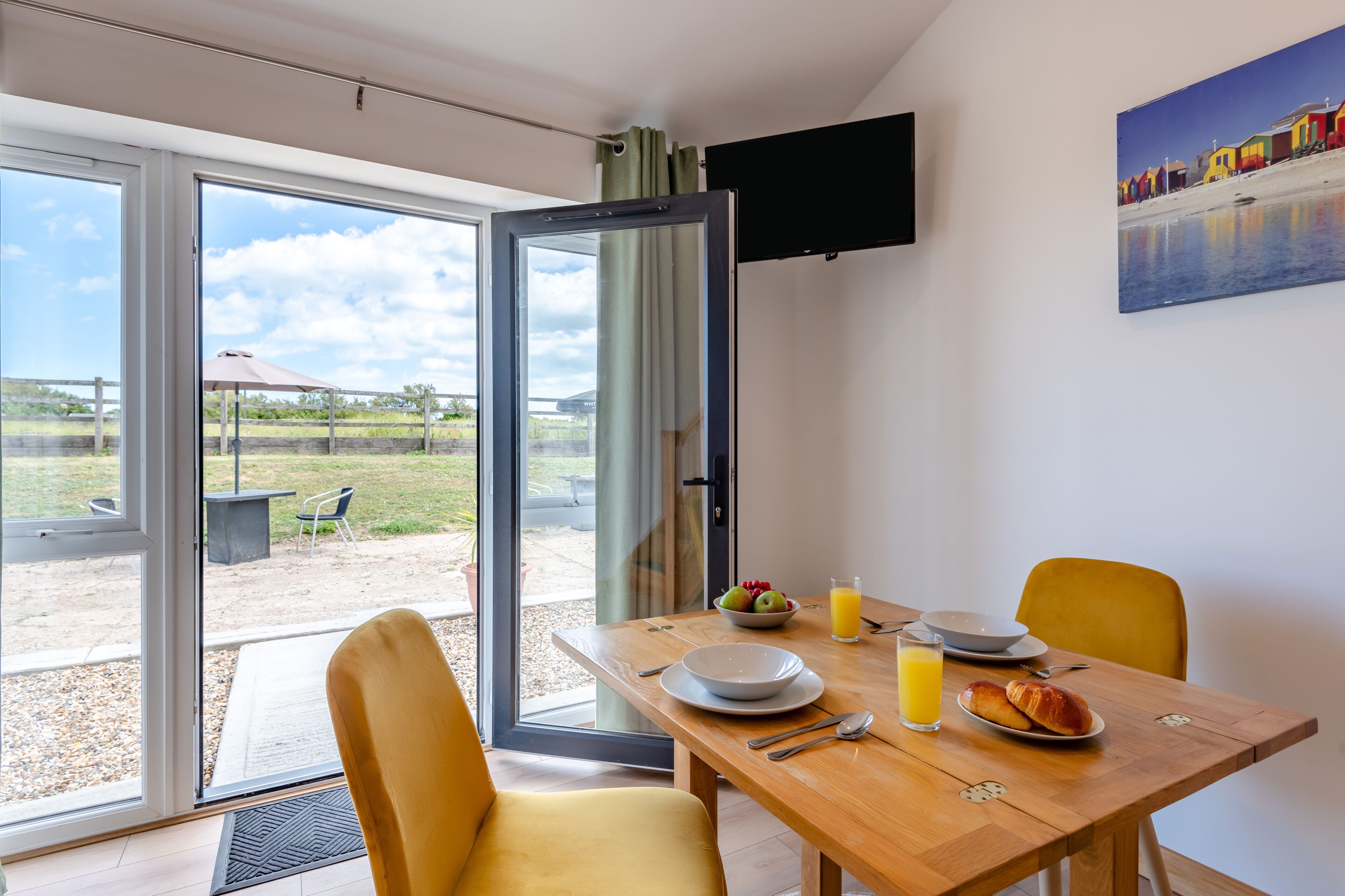 Un comedor con una mesa preparada para dos, un televisor montado en la pared y vistas al exterior a través de la puerta de cristal.