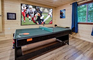 Une table de ping-pong dans une pièce aux murs en bois, un téléviseur diffusant un match de football et une fenêtre avec des rideaux.