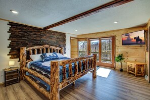 Un lit avec un cadre en bois, un mur d’accent en pierre, une grande fenêtre offrant une belle vue et un téléviseur fixé au mur.