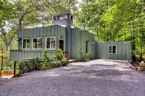 Une cabane en bois peinte en vert, dotée de nombreuses fenêtres, d’une cheminée et d’une allée de gravier, le tout entouré d’arbres.