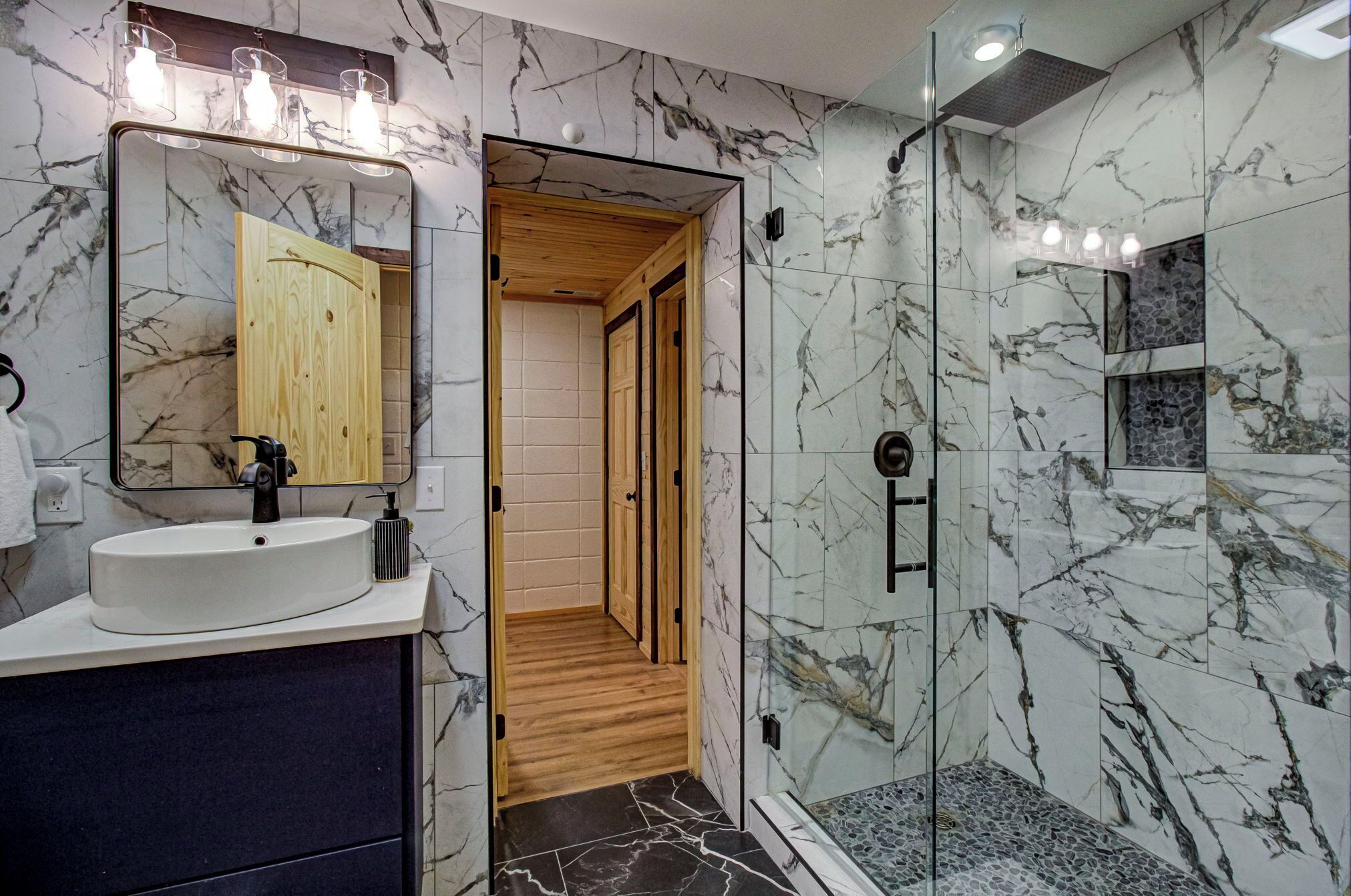 A modern bathroom with a marble backsplash, a wooden vanity, a glass shower enclosure, and a large mirror.