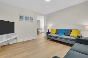 A modern living room with a flat-screen TV, a grey sofa with yellow cushions, and a wooden dining table. - Newly Renovated - Pet OK - 2BR Steps to Beach (Wildwood)