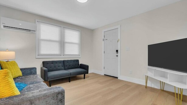 Ein Wohnzimmer mit grauem Sofa, einem Fernseher auf einem weißen Schrank und einem Fenster mit Jalousien.