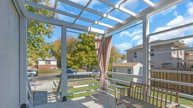 Eine überdachte Terrasse mit Blick auf eine Wohnstraße, einer Holzbank und einem Tisch mit einer Flasche.