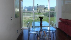 Um interior moderno com uma mesa de jantar e cadeiras, um sofá vermelho e amplas janelas com vista para edifícios e áreas verdes.