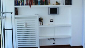 Una habitación con una estantería blanca, un televisor montado en la pared y una ventana con persianas.