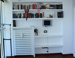 Um quarto com uma estante branca, uma televisão fixada na parede e uma janela com persianas.