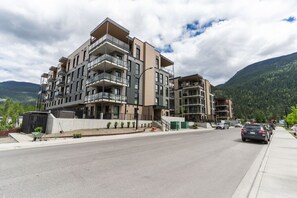 Un immeuble résidentiel à plusieurs étages, doté de balcons, situé le long d’une route pavée, avec une chaîne de montagnes en arrière-plan.