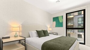Habitación de hotel con cama, mesitas de noche, lámpara, ventana con vistas a edificios y un cuadro en la pared.