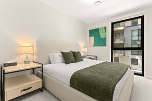 Ein Hotelzimmer mit Bett, Nachttischen, einer Lampe, einem Fenster mit Blick auf Gebäude und einem Bild an der Wand.