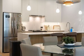 Eine moderne Küche mit Edelstahlgeräten, einer zentralen Kochinsel mit Sitzgelegenheiten, Pendelleuchten und einem sichtbaren Mülleimer.