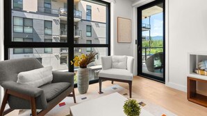 Una sala de estar moderna con un sofá, un sillón y una mesa de centro. Hay un ventanal con vistas a un edificio y un balcón.
