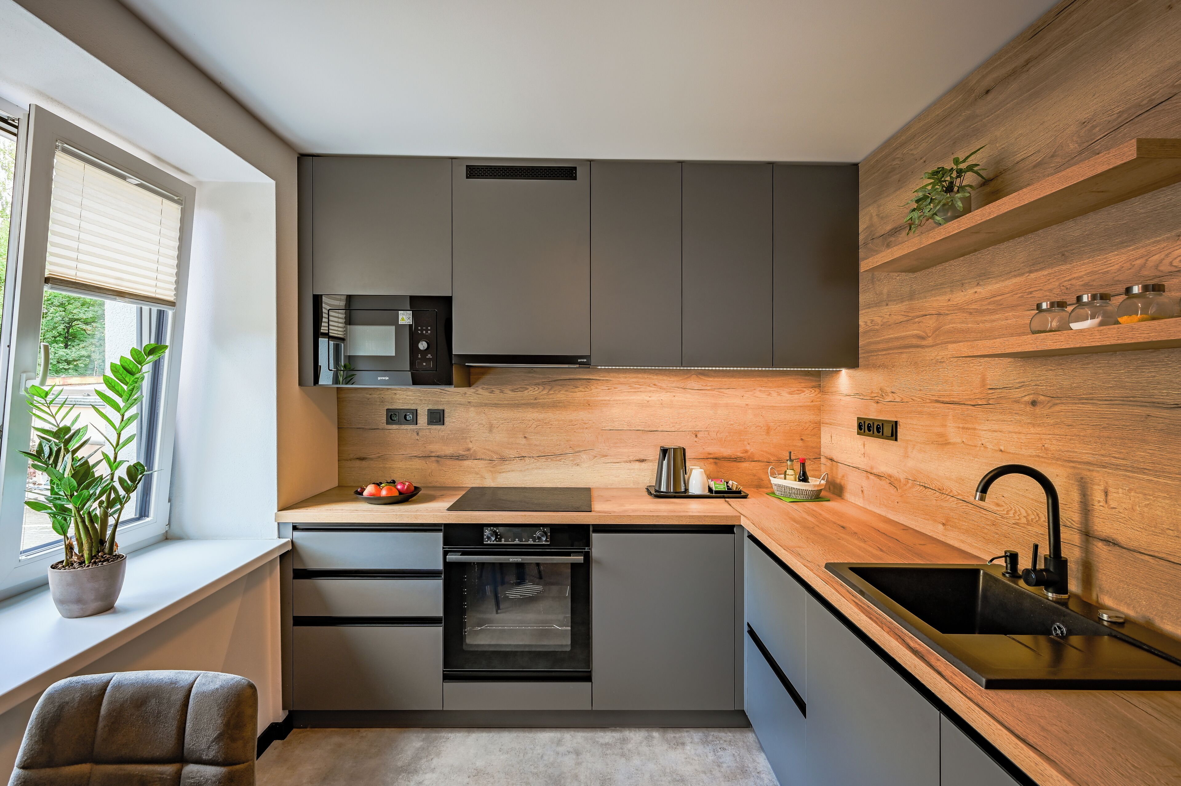 Cocina moderna con armarios grises, horno empotrado, microondas y una pared de acento de madera.