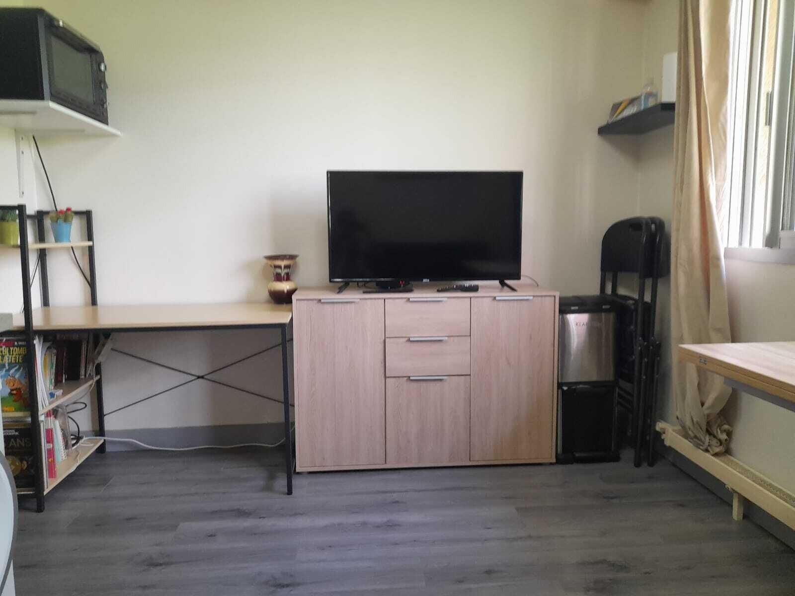 Een knus woongedeelte met een flatscreen televisie op een houten kast, een boekenkast met boeken, een klein tafeltje en een kitchenette met een magnetron en waterkoker.