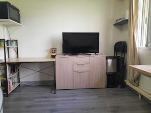 Een knus woongedeelte met een flatscreen televisie op een houten kast, een boekenkast met boeken, een klein tafeltje en een kitchenette met een magnetron en waterkoker.