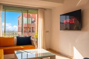 A modern living room with a sofa, a glass coffee table, and a large window offering a view of buildings. A flat-screen TV mounted on the wall displays the Netflix logo. - Hivernage Luxury Pool and Balcony (Marrakech)