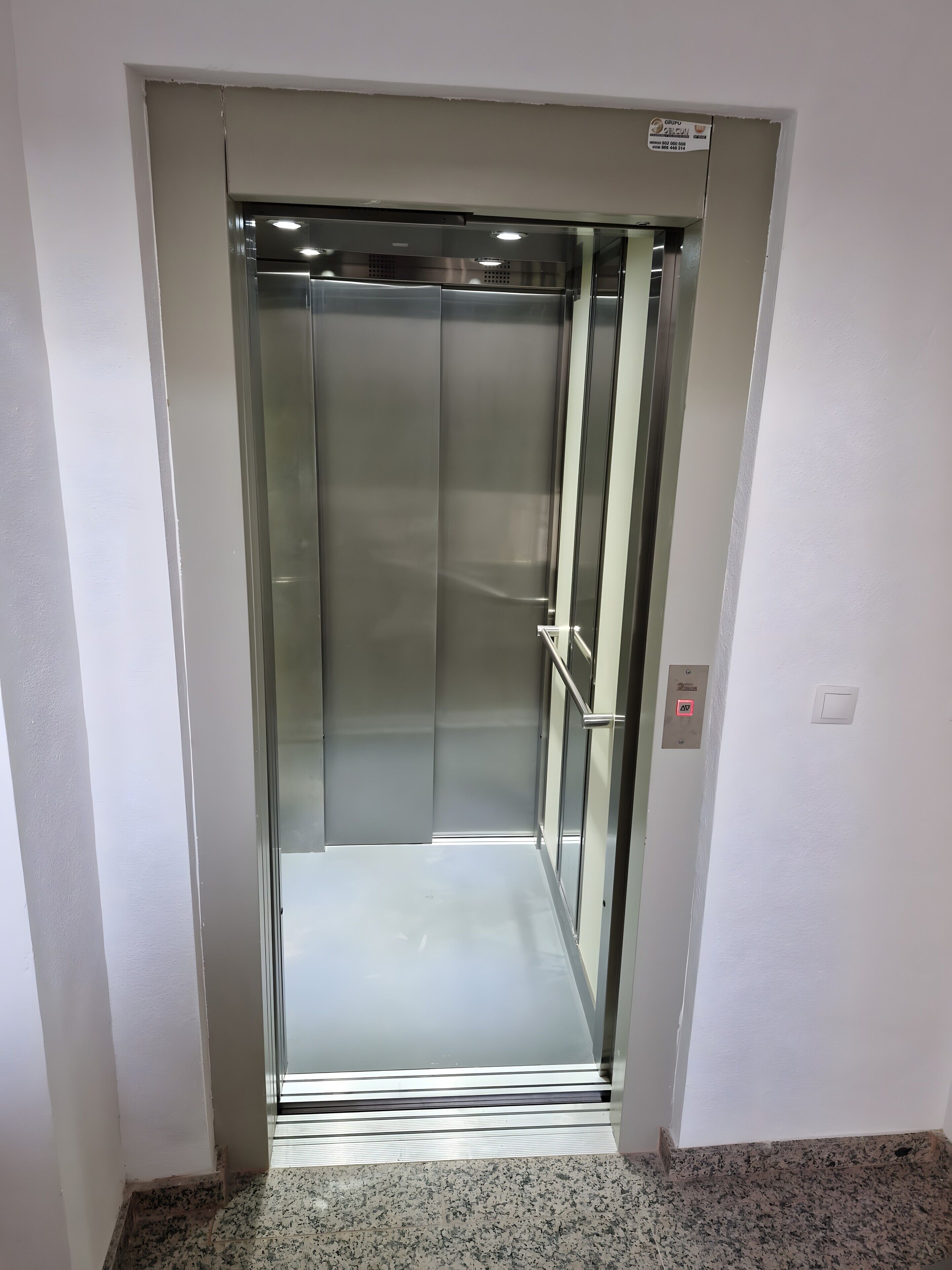 Ein Aufzug mit metallischer Innenausstattung und Glastür, ausgestattet mit Bedienfeld und Notrufknopf.