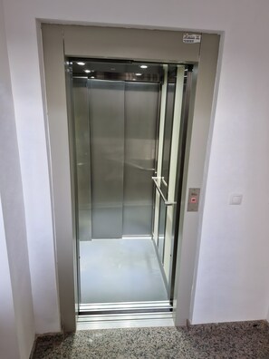 Een lift met een metalen inrichting en een glazen deur, met een bedieningspaneel en een noodknop.