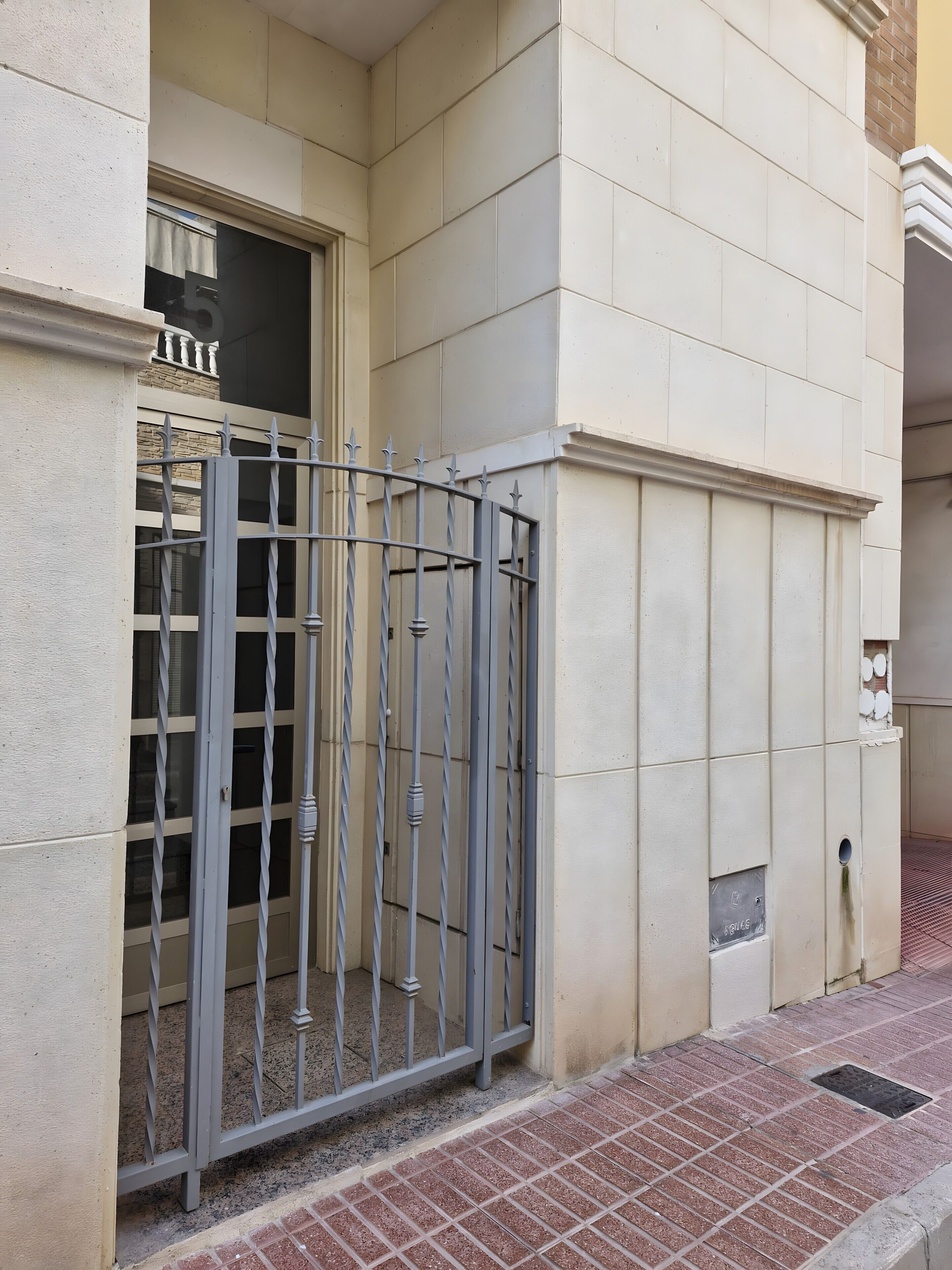 Une façade d’immeuble avec un portail en métal, une fenêtre avec un rideau et un sol carrelé.