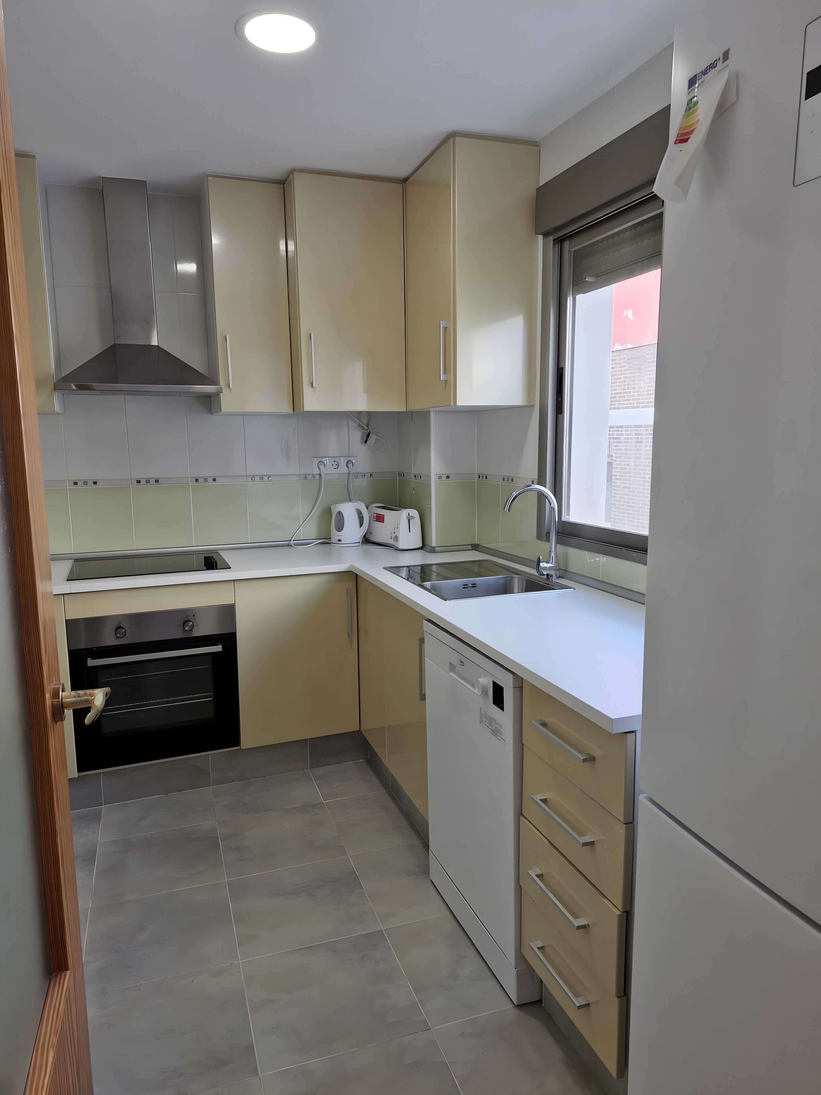 A kitchen with beige cabinets, a stainless steel range hood, a dishwasher, and a window with a view.