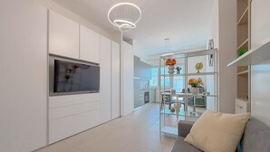 A modern kitchen with white cabinetry, a built-in TV, and a dining area with a glass table and chairs. - Bright Studio with A/C and Balcony (Civitanova Marche)