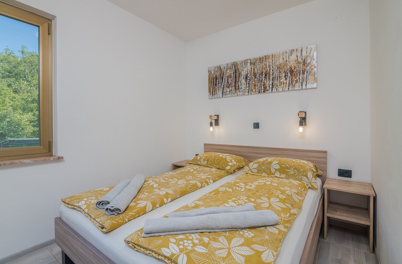 A double bed with floral bedding, a bedside table, a wall-mounted light, and a window with a view of trees.